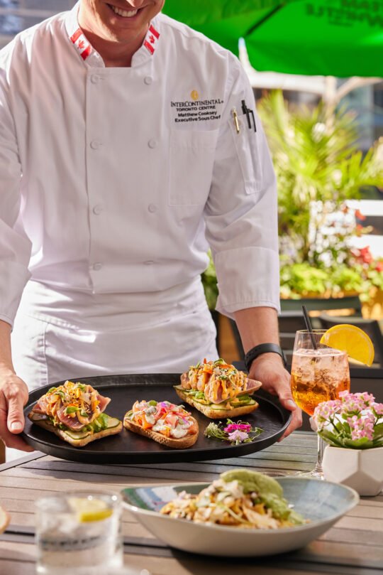 A chef bringing a beautifully prepared dish to the table, ready to be served in a warm outdoor summer setting. Natural light, fresh atmosphere, and thoughtful composition capture the feeling of a perfect dining moment.