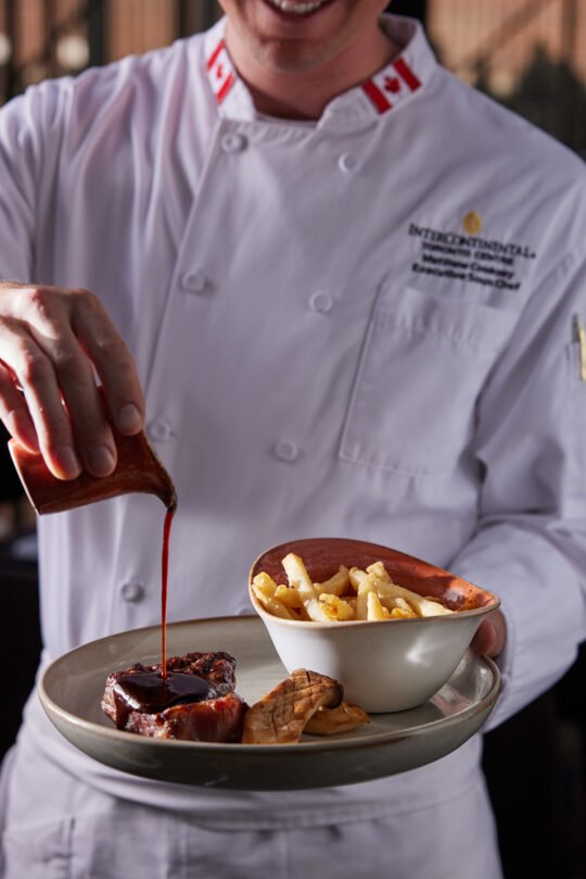 A chef pouring the sauce to a dish ready to be served in a dark mood setting. Natural light, moody atmosphere capture the feeling of a perfect dining moment.
