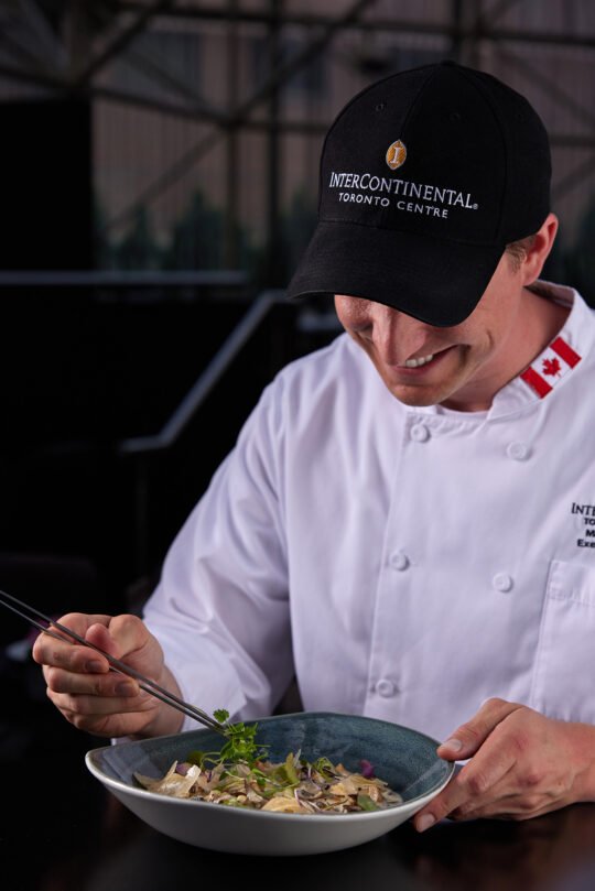 A chef garnishing and prepared a beautifully prepared dish on the table ready to be served in a dark mood setting. Natural light, moody atmosphere capture the feeling of a perfect dining moment.