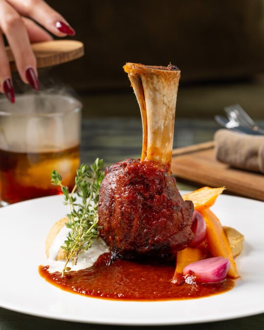 Close up photography of lamb steak, behind a hand open a smoke cocktail blue in the background