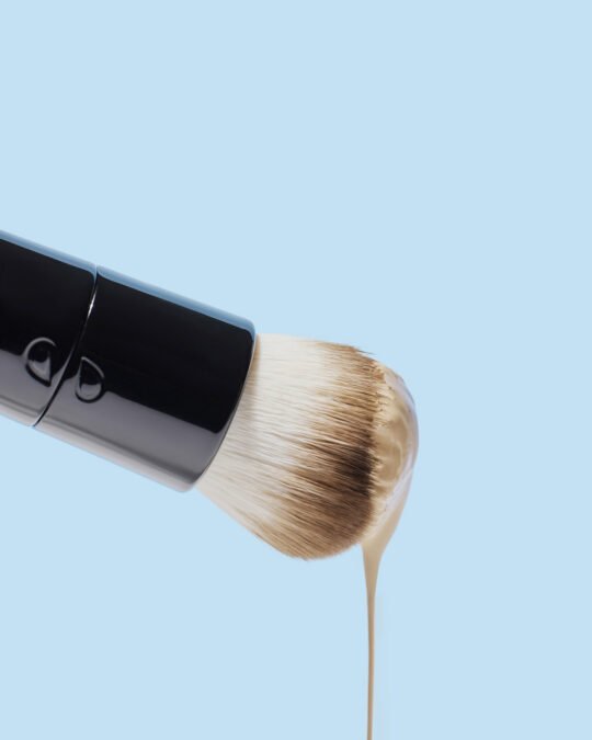 Commercial beauty product photography of a make up brush with liquid on the tip falling on blue background