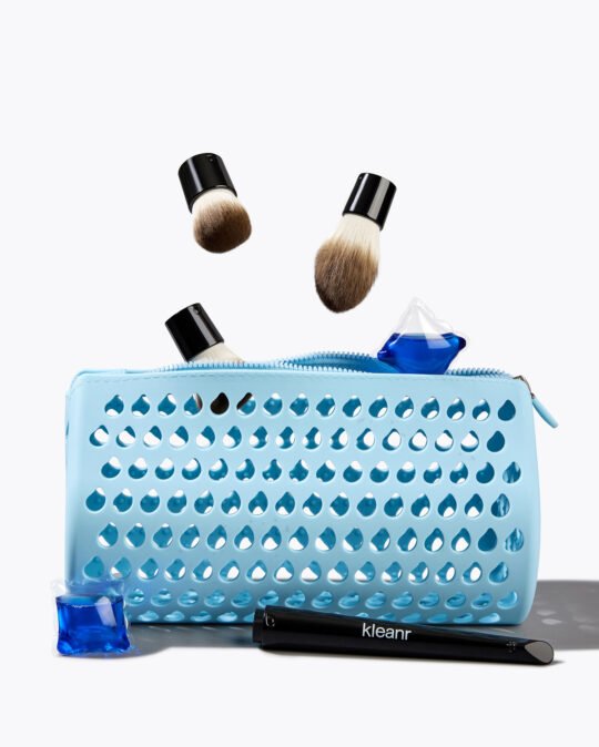 Commercial product skincare photography of a blue washing bag with floating makeup brushes bristles and pods on a white background.