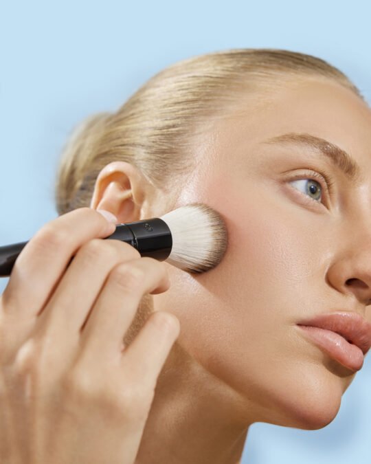 Professional beauty photography of a model applying makeup with a foundation brush, showcasing flawless skin for a cosmetics brand campaign in Toronto