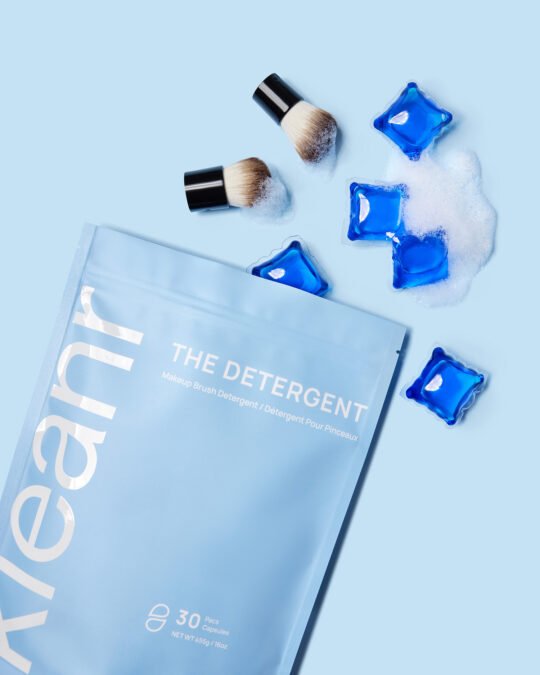 Product Photography of detergent pouch laying on the blue background with pods and make up brush's bristles surround it, bright mood tone