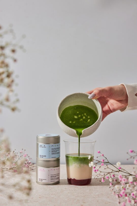 Photoshoot of pouring a matcha to a glass to make a matcha strawberry, in the soft light with some small flower in the foreground blurry.