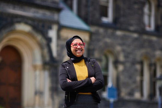 Woman in dark coat standing on University of Toronto campus, natural light historic building in background.