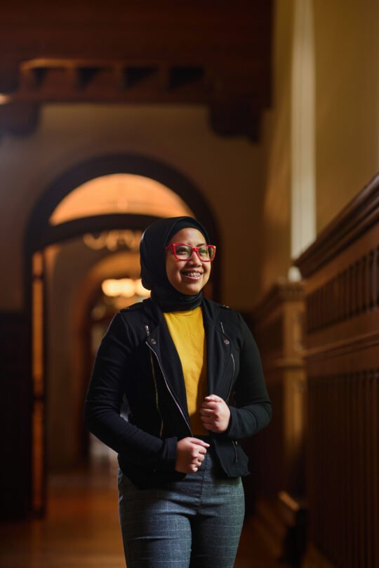 Portrait of woman in dark coat standing inside University of Toronto campus, moody lighting and historic building in background.